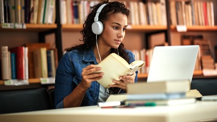 Junge Frau sitzt mit Kopfhörern und einem Buch an einem Tisch in der Bibliothek und lernt.