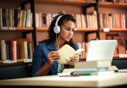Junge Frau sitzt mit Kopfhörern und einem Buch an einem Tisch in der Bibliothek und lernt.