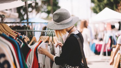 Frau auf einem Flohmarkt stöbert in gebrauchten Kleidungsstücken, die auf einem Kleiderständer hängen.