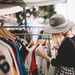 Frau auf einem Flohmarkt stöbert in gebrauchten Kleidungsstücken, die auf einem Kleiderständer hängen.