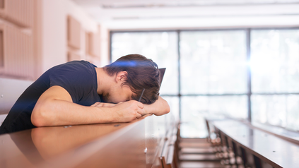 Erschöpfter Student in einem Hörsaal veschränkt die Arme auf dem Tisch vor sich und stützt seinen Kopf darauf.