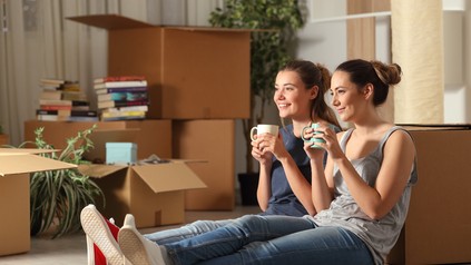 Zwei Studentinnen sitzen zwischen Umzugskartons auf dem Boden in ihrem neuen WG-Wohnzimmer.
