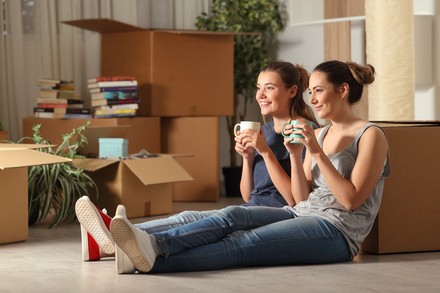 Zwei Studentinnen sitzen zwischen Umzugskartons auf dem Boden in ihrem neuen WG-Wohnzimmer.