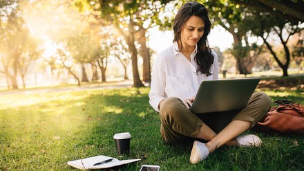 eine junge Frau sitzt in einem Park mit einem Laptop, Kaffee und einen Buch zum schreiben.