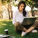 eine junge Frau sitzt in einem Park mit einem Laptop, Kaffee und einen Buch zum schreiben.