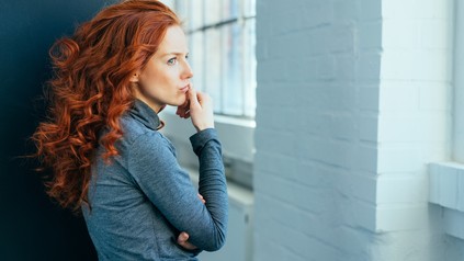 Frau mit langen roten Locken schaut traurig aus dem Fenster.