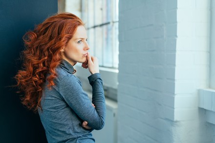 Frau mit langen roten Locken schaut traurig aus dem Fenster.