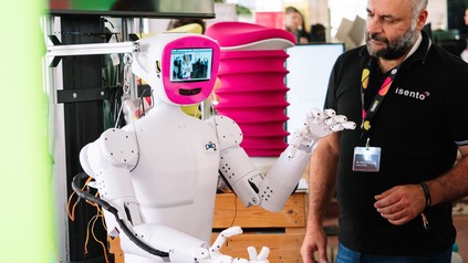 Ein weißer Roboter mit einem kleinen Bildschirm auf der Brust interagiert mit einem Messebesucher.