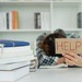 Studentin am Schreibtisch hält einen Zettel mit dem Wort "Help" in die Luft und hat ihren Kopf in das Buch gelegt.