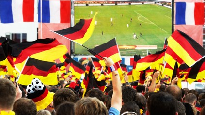 Viele menschen in Fanbekleidung und mit Deutschland-Fähnchen schauen gemeinsam ein match auf Großbildleinwand.