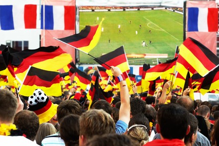 Viele menschen in Fanbekleidung und mit Deutschland-Fähnchen schauen gemeinsam ein match auf Großbildleinwand.