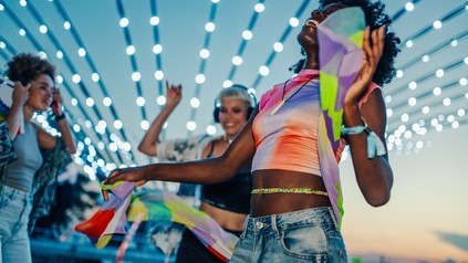 Drei Raverinnen tanzen auf einer Open-Air-Veranstaltung unter Lichterketten.