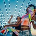 Drei Raverinnen tanzen auf einer Open-Air-Veranstaltung unter Lichterketten.