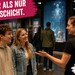 Junge Teammitarbeiterin spricht in einer modernen Ausstellung mit zwei Besucher:innen. Text im Bild wirbt für Studentenjobs bei Events.