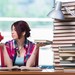 Junge Frau hält in der einen Hand ein Sparschein und in der anderen Hand ein Buch. Sie blickt sorgenvoll auf das Sparschwein.