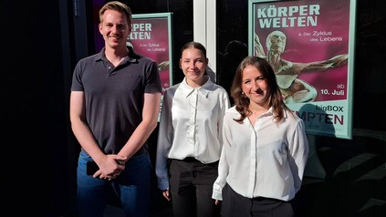 Zwei junge Frauen und ein Mann vor einem Plakat der Ausstellung KÖRPERWELTEN