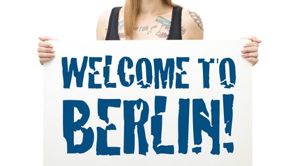 Junge Berlinerin mit Sonnenbrille und Mütze hält Schild mit der Aufschrift "Welcome to Berlin" in die Kamera.