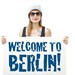 Junge Berlinerin mit Sonnenbrille und Mütze hält Schild mit der Aufschrift "Welcome to Berlin" in die Kamera.