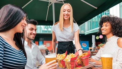 Außengastronomie, drei junge Gäste sitzen vor Getränken, junge Bedienung bringt ein Tablett mit Pommes.