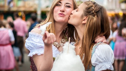 Zwei Studentinnen in Dirndl-Kleidern essen Zuckerwatte auf dem Oktoberfest.