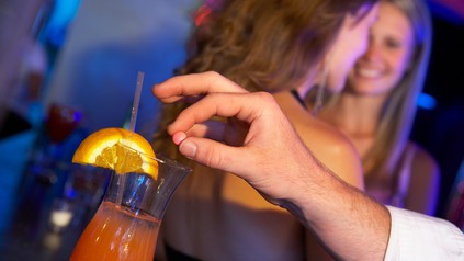 Männerhand wirft heimlich eine Pille in ein Getränk; im Hintergrund feiern zwei junge Frauen auf einer Tanzfläche.