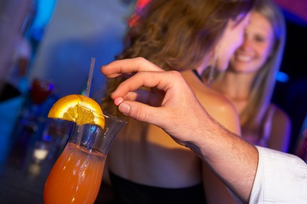 Männerhand wirft heimlich eine Pille in ein Getränk; im Hintergrund feiern zwei junge Frauen auf einer Tanzfläche.