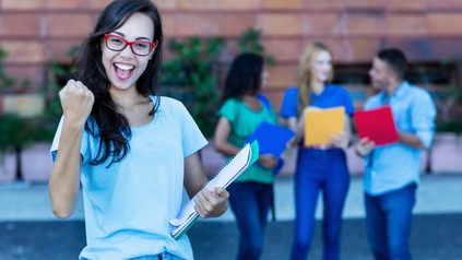 Junge Studentin jubelt mit Unterlagen in der Hand vor einer Hochschule, im Hintergrund stehen weitere Studierende.