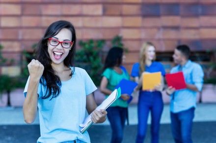 Junge Studentin jubelt mit Unterlagen in der Hand vor einer Hochschule, im Hintergrund stehen weitere Studierende.