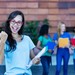 Junge Studentin jubelt mit Unterlagen in der Hand vor einer Hochschule, im Hintergrund stehen weitere Studierende.