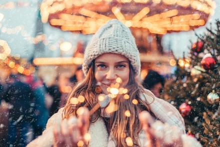 Junge Frau mit Wollmütze hält brennende Wunderkerzen auf einem weihnachtlich beleuchteten Weihnachtsmarkt.