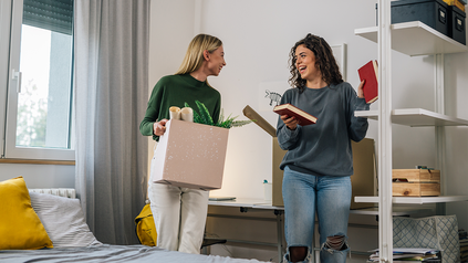 Zwei junge Frauen richten ein kleines Zimmer ein und unterhalten sich dabei. Eine Frau trägt eine Karton mit Pflanzen, die andere hält ein Buch in der Hand.
