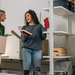 Zwei junge Frauen richten ein kleines Zimmer ein und unterhalten sich dabei. Eine Frau trägt eine Karton mit Pflanzen, die andere hält ein Buch in der Hand.