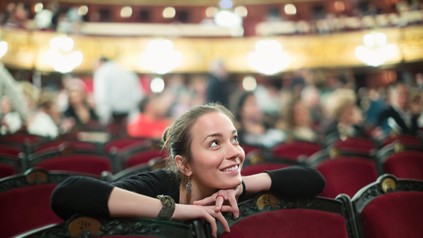 Junge Frau sitzt in einem Theater mit roten Sesseln und blickt lächelnd zur Bühne.