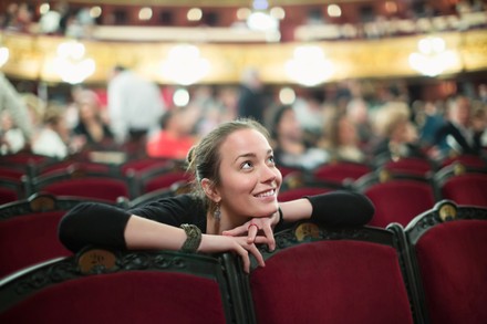Junge Frau sitzt in einem Theater mit roten Sesseln und blickt lächelnd zur Bühne.