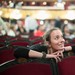 Junge Frau sitzt in einem Theater mit roten Sesseln und blickt lächelnd zur Bühne.