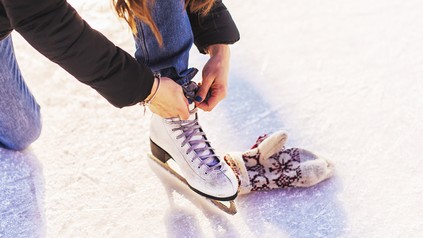Person schnürt einen weißen Schlittschuh auf einer Eisfläche. Daneben liegt ein gemusterter Winterhandschuh.