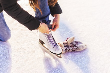 Person schnürt einen weißen Schlittschuh auf einer Eisfläche. Daneben liegt ein gemusterter Winterhandschuh.