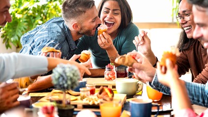 Junge Erwachsene sitzen lachend an einem gedeckten Frühstückstisch mit Gebäck, Kaffee und Orangensaft.