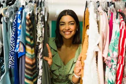 Lächelnde junge Frau in einem grünen Kleid stöbert zwischen dicht behängten Kleiderständern in einem Secondhand-Laden.