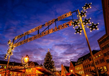 Beleuchteter Schriftzug „Willkommen auf dem Christkindlesmarkt“ über festlich geschmückten Weihnachtsmarktständen bei Abenddämmerung.