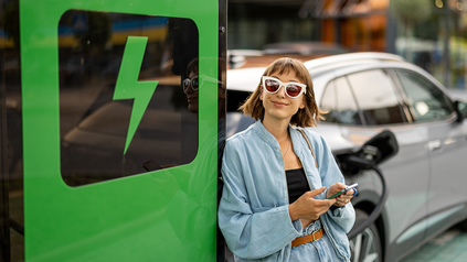 Junge Dame mit Smartphone an der Hand lehnt an E-Ladesäule, während ihr Auto lädt.