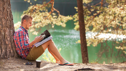 Junger Mann lehnt an Baum und liest Magazin.